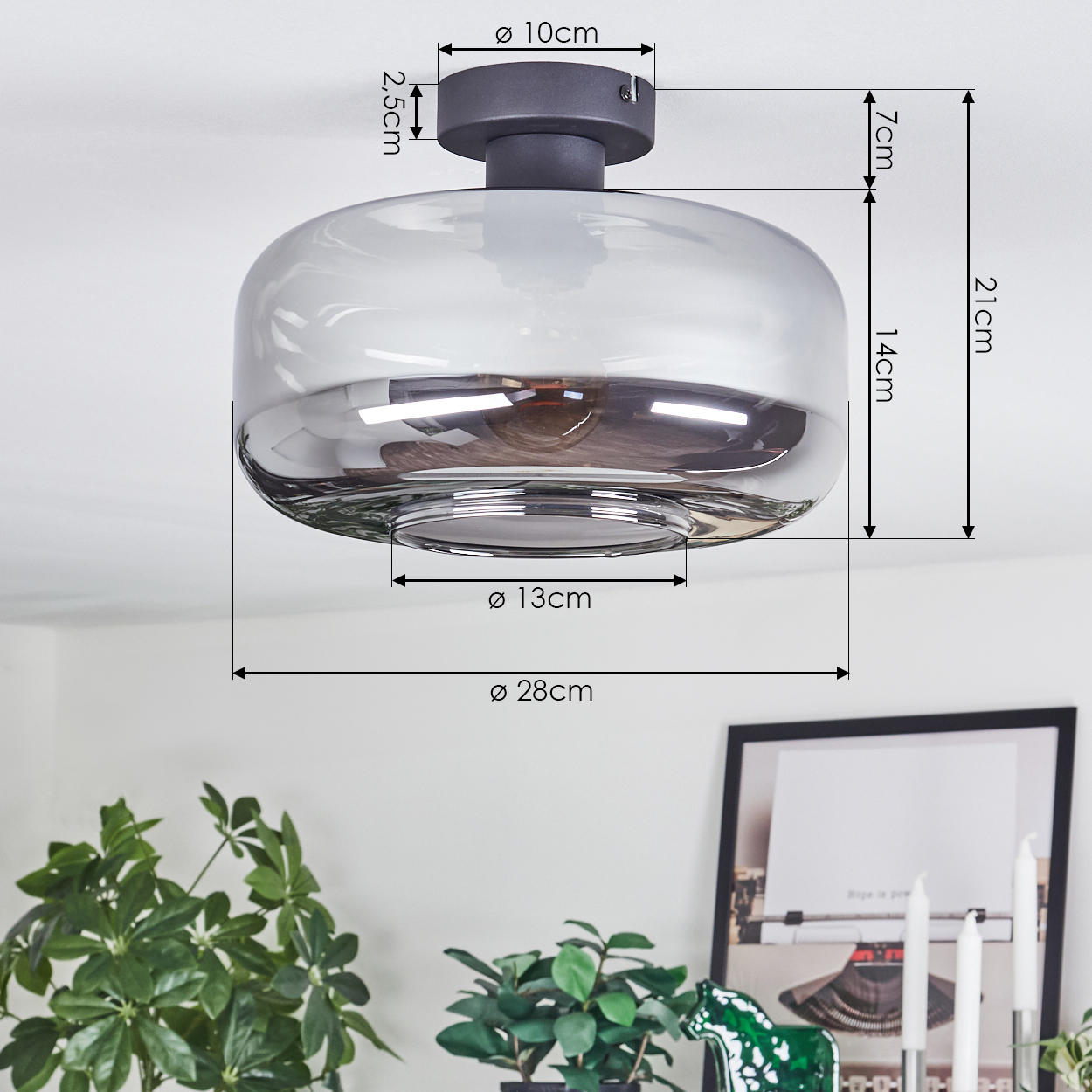 Eine moderne Deckenlampe mit Maßen wird angezeigt. Im Vordergrund sind grüne Pflanzen zu sehen, im Hintergrund ein gerahmtes Bild.