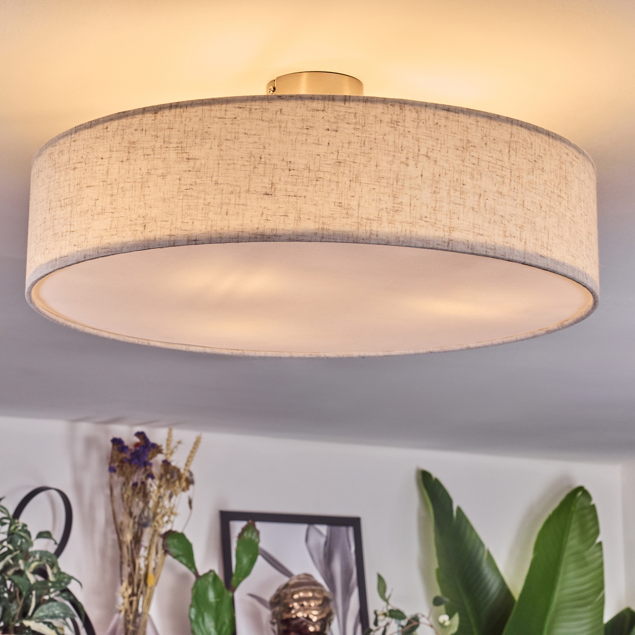 Eine runde Deckenlampe mit Stoffschirm hängt über Pflanzen und einem Bilderrahmen. Die Lampe ist beige mit grauem Rand.