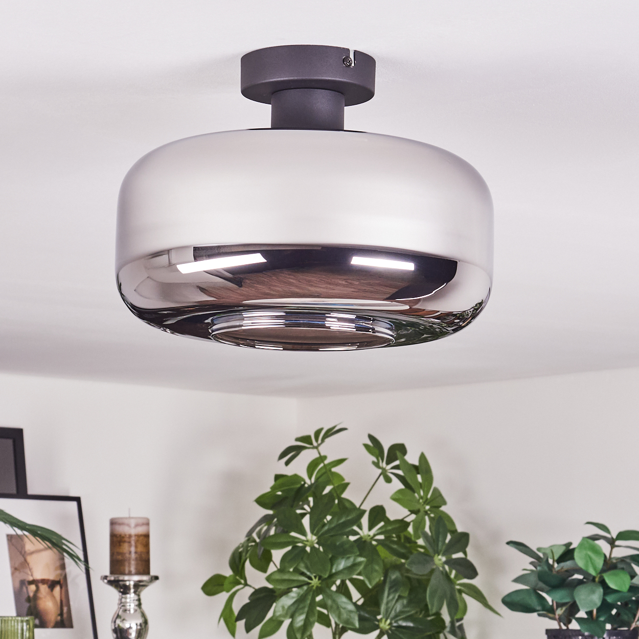 Eine moderne, glänzende Deckenlampe. Darunter sind Pflanzen und Dekoration zu sehen. Die Lampe reflektiert den Raum. Der Hintergrund ist weiß.