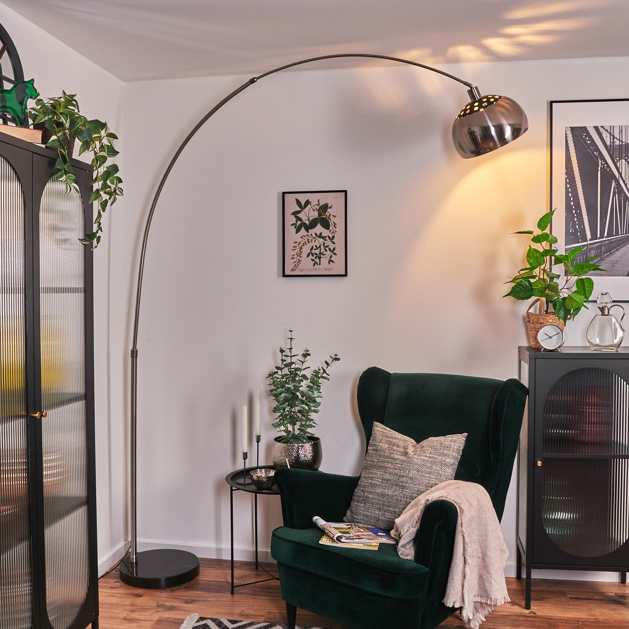 Wohnzimmer mit grünem Sessel, Stehlampe und schwarzem Schrank. Pflanzen und gerahmte Kunst sind ebenfalls sichtbar.
