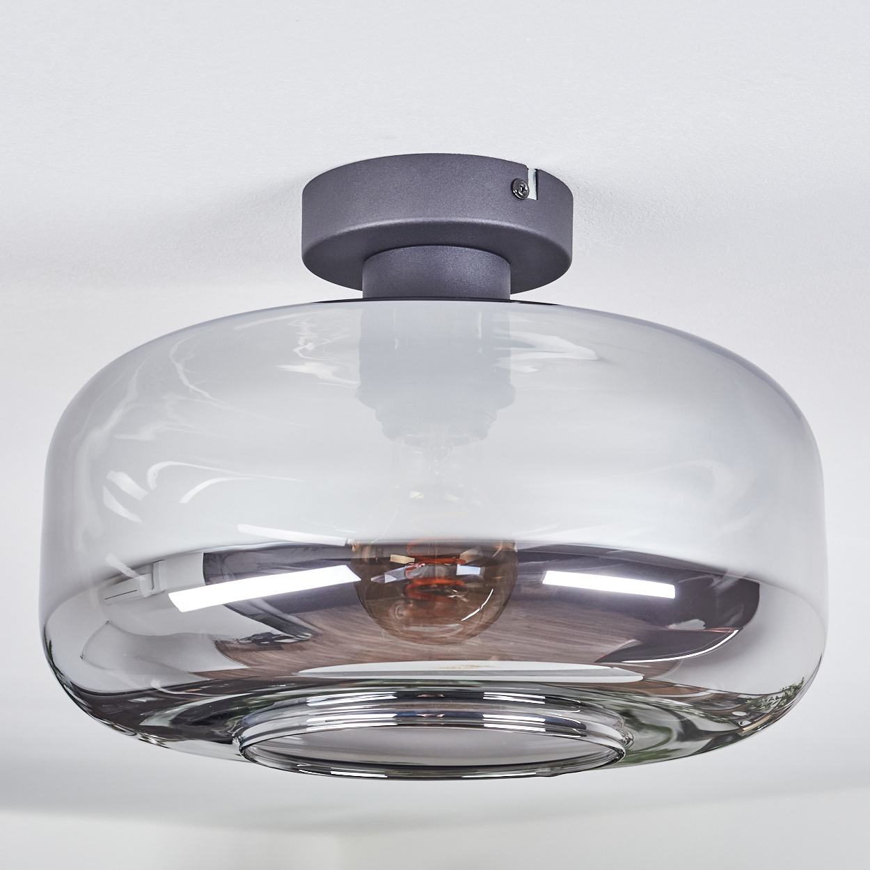 Eine moderne Deckenlampe aus Rauchglas hängt von einer weißen Decke. Die Lampe hat einen dunkelgrauen Sockel und einen runden, transparenten Schirm.