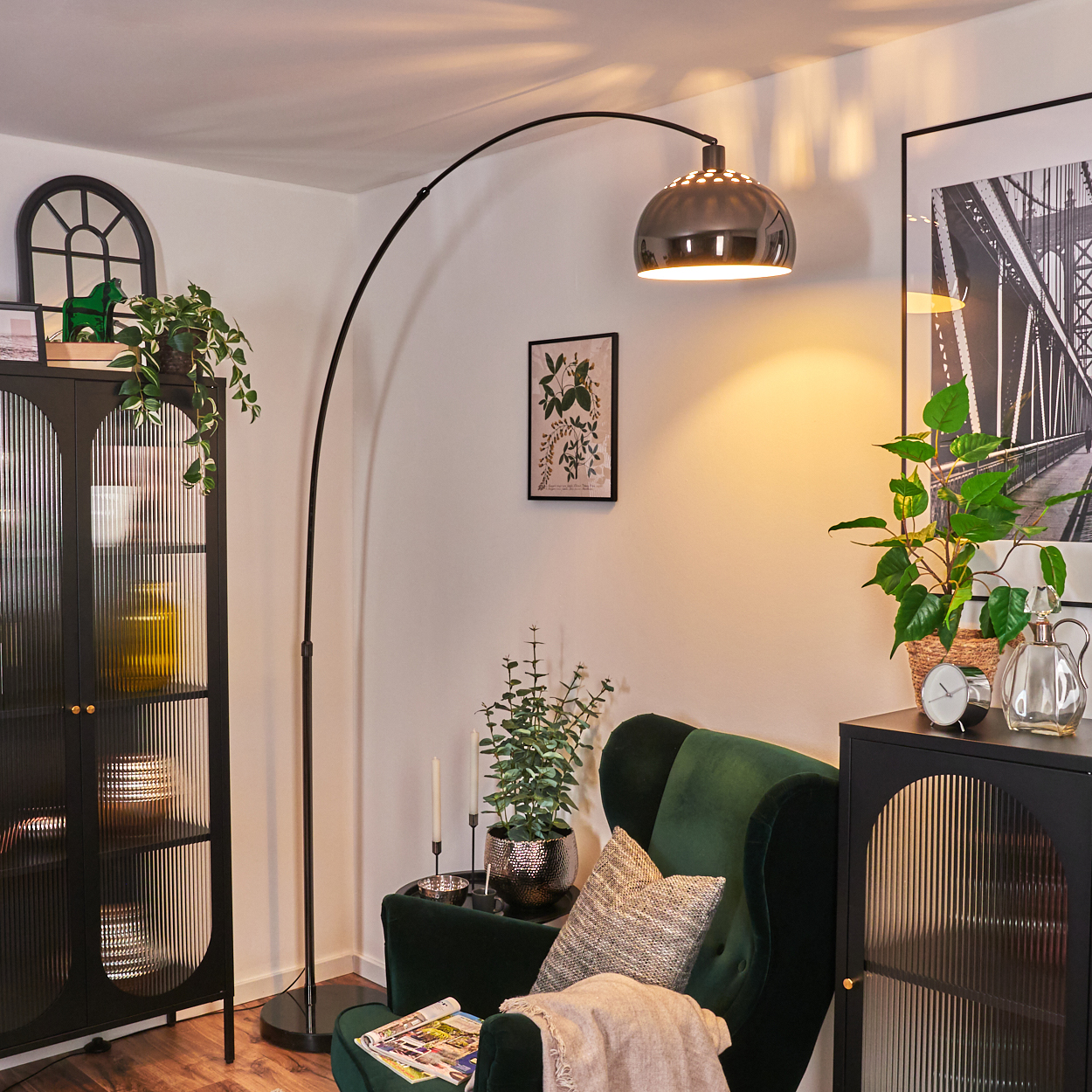 Ein modernes Wohnzimmer mit grünem Sessel, Stehlampe, Pflanzen und einem schwarzen Schrank. Ein Brückenfoto hängt an der Wand.