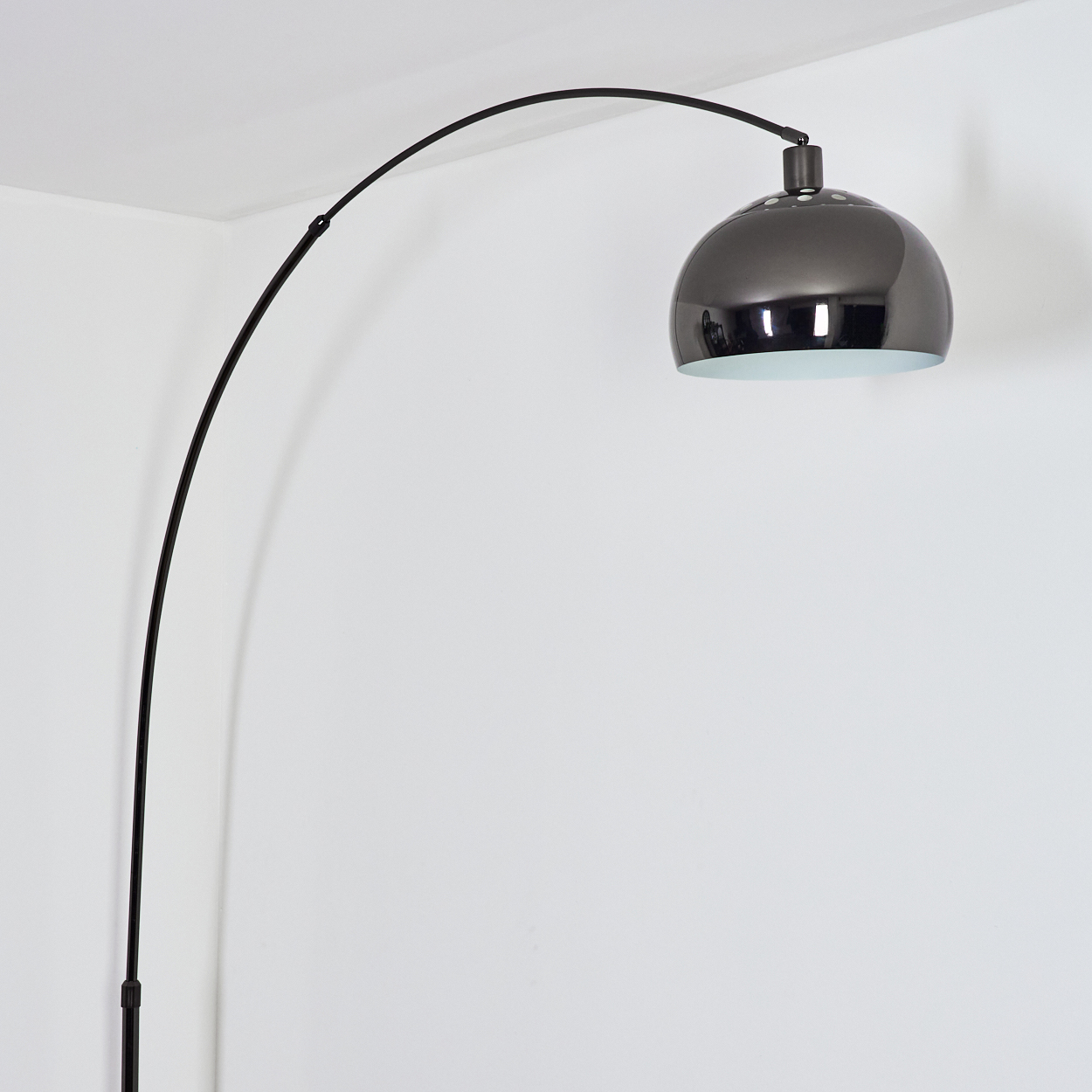 Eine moderne Bogenlampe mit schwarzem, gebogenem Arm und dunklem, metallischem Lampenschirm, vor einer weißen Wand.