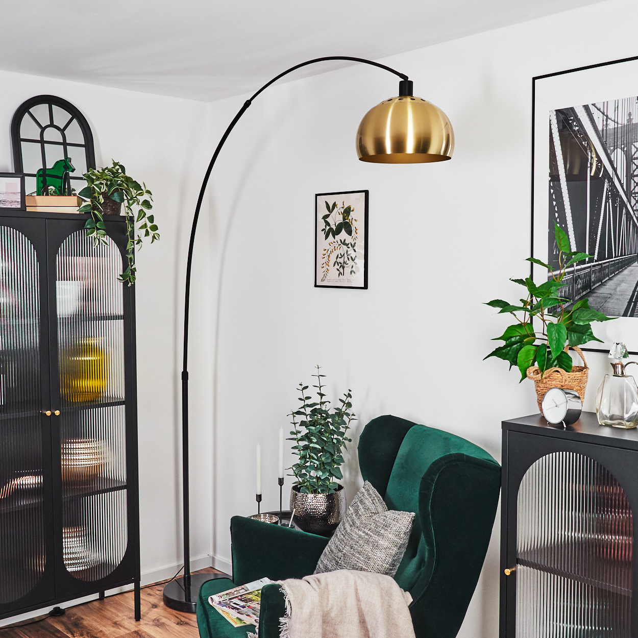 Ein Wohnzimmer mit grünem Sessel, Stehlampe, schwarzem Schrank und gerahmter Kunst. Pflanzen und Dekorartikel ergänzen die Einrichtung.