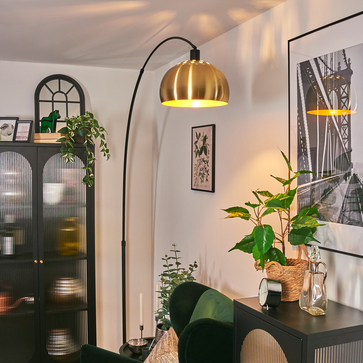 Ein Raum mit einer goldenen Lampe, einem grünen Sofa, einem schwarzen Schrank und einem gerahmten Brückenfoto. Pflanzen und Dekorationen sind ebenfalls vorhanden.