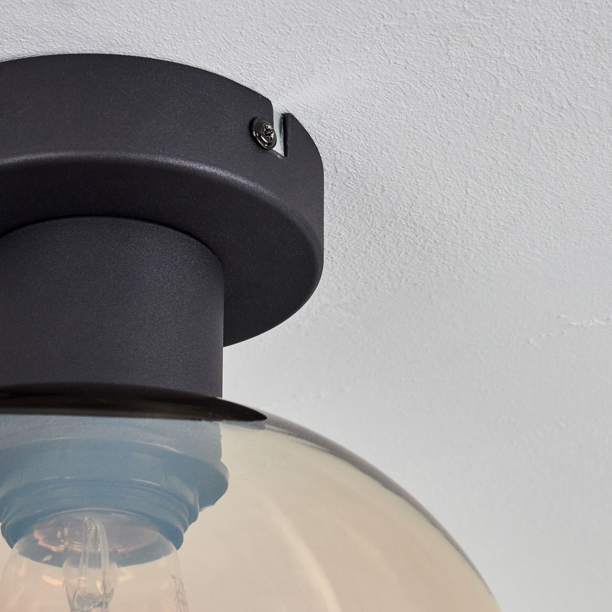 Nahaufnahme einer schwarzen Deckenlampe. Ein Glasschirm ist sichtbar. Die Lampe ist an einer strukturierten weißen Decke montiert.