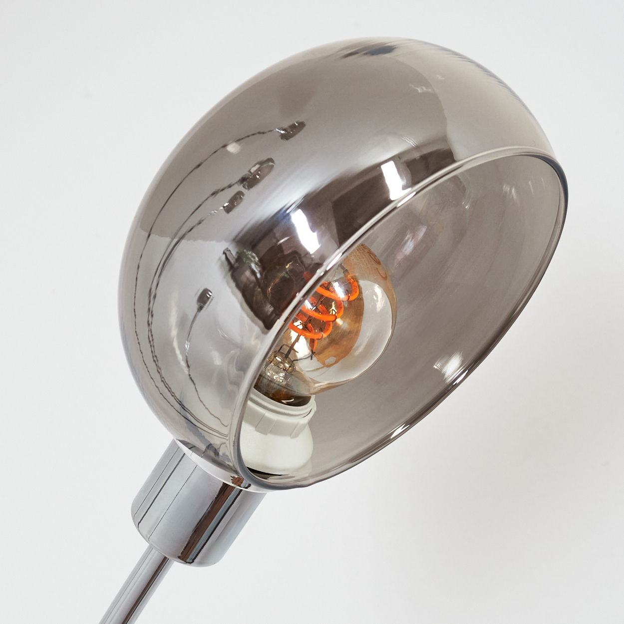 Nahaufnahme einer modernen Lampe. Ein Chromschaft trägt einen rauchigen Glasschirm. Im Inneren ist eine leuchtende orangefarbene Glühbirne zu sehen.