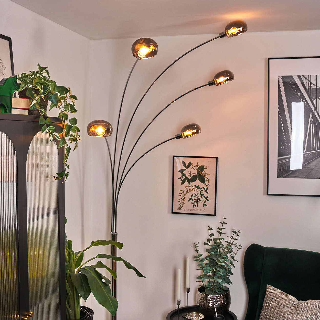Ein modernes Wohnzimmer mit Stehlampe, Pflanzen und gerahmter Kunst. Die Lampe hat fünf Lichter. Links ein schwarzer Schrank.