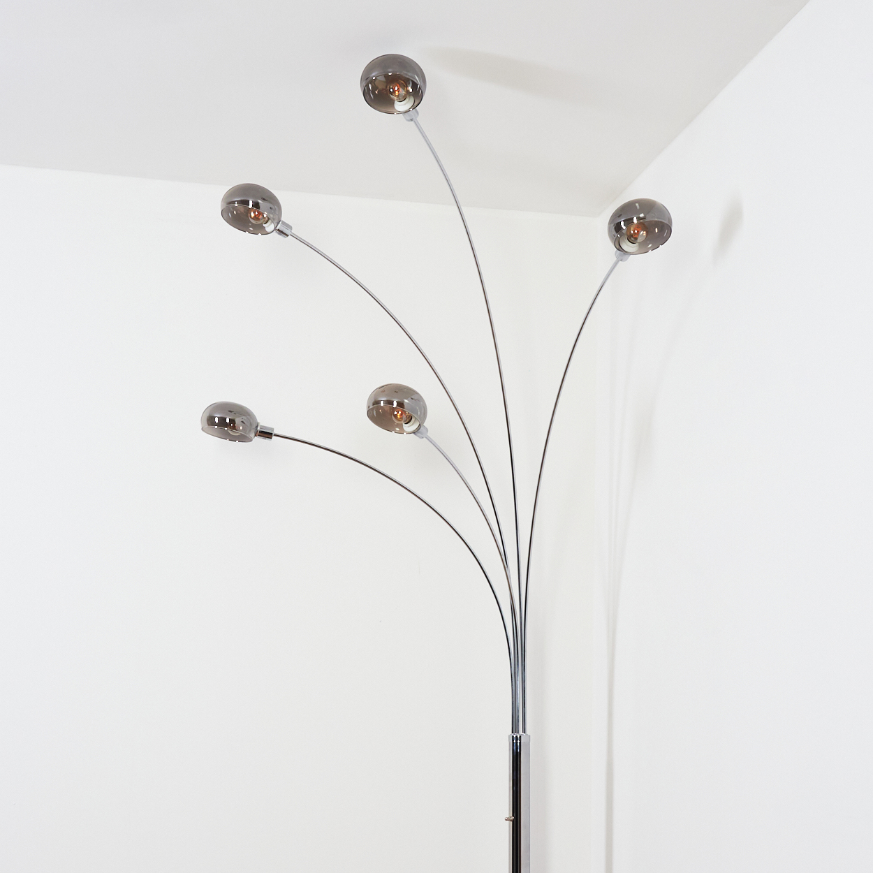 Eine moderne Stehlampe mit fünf gebogenen Armen, die jeweils einen rauchigen Glaskugelschirm tragen. Die Lampe ist verchromt, vor einer weißen Wand.