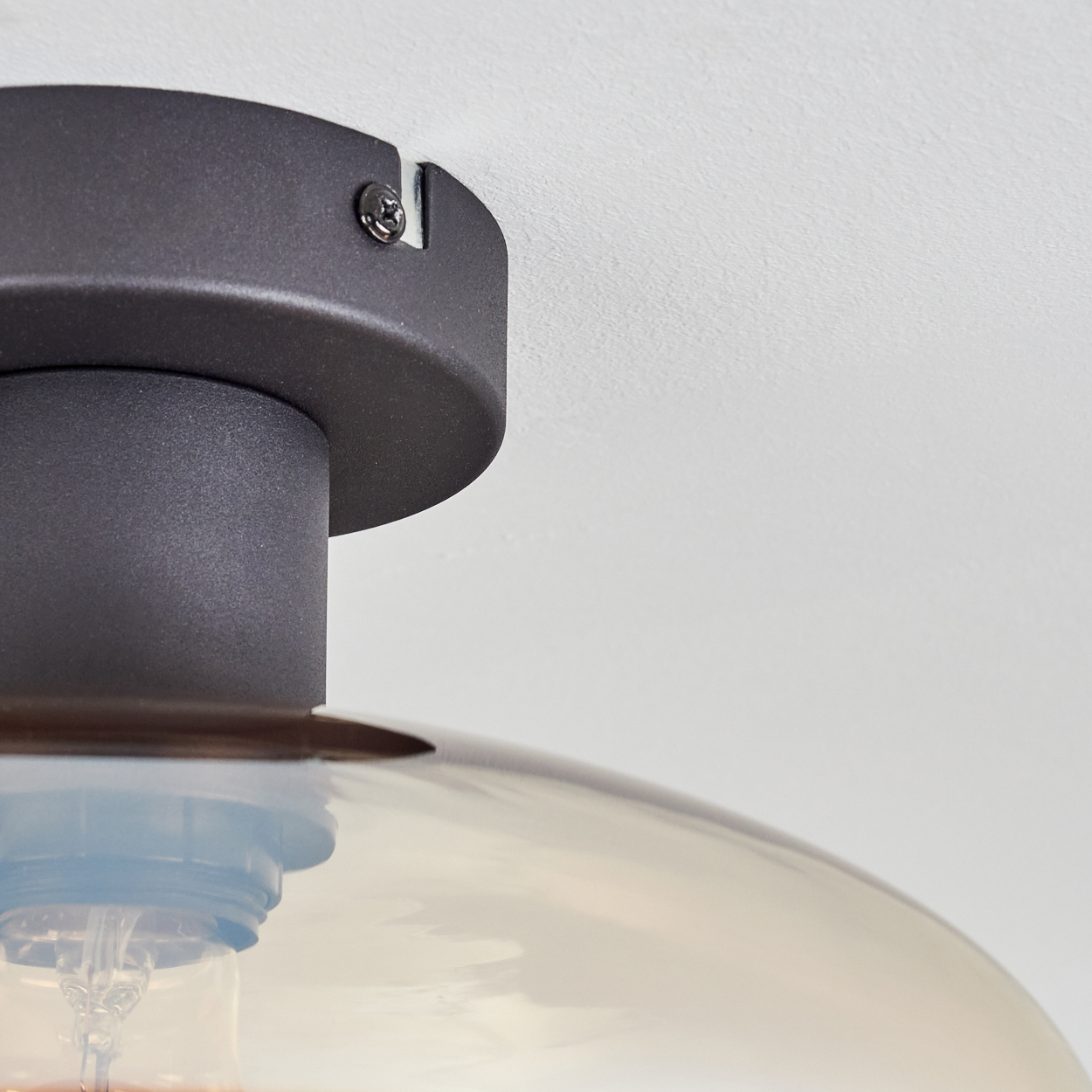 Nahaufnahme einer Deckenleuchte. Ein dunkler Metallsockel ist an der weißen Decke befestigt. Die Leuchte hat einen Glasschirm.