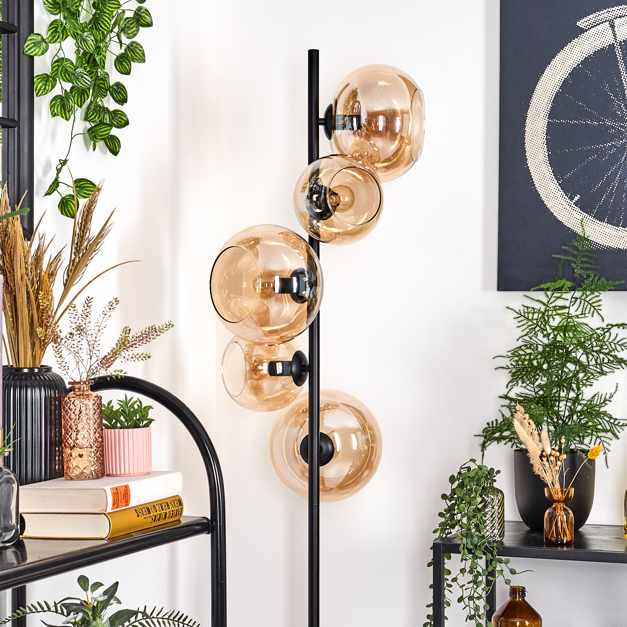 Eine moderne Stehlampe mit fünf bernsteinfarbenen Glaskugeln. Verschiedene Pflanzen und Dekorationen umgeben sie. Ein Fahrradrad-Kunstwerk hängt im Hintergrund.