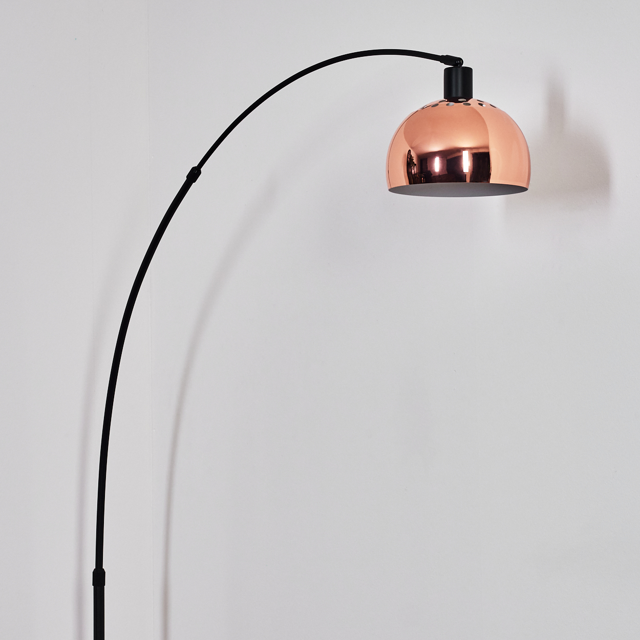 Eine moderne Bogen-Stehlampe mit kupferfarbenem Lampenschirm vor einer weißen Wand. Die Lampe hat einen schwarzen, gebogenen Arm und Fuß.