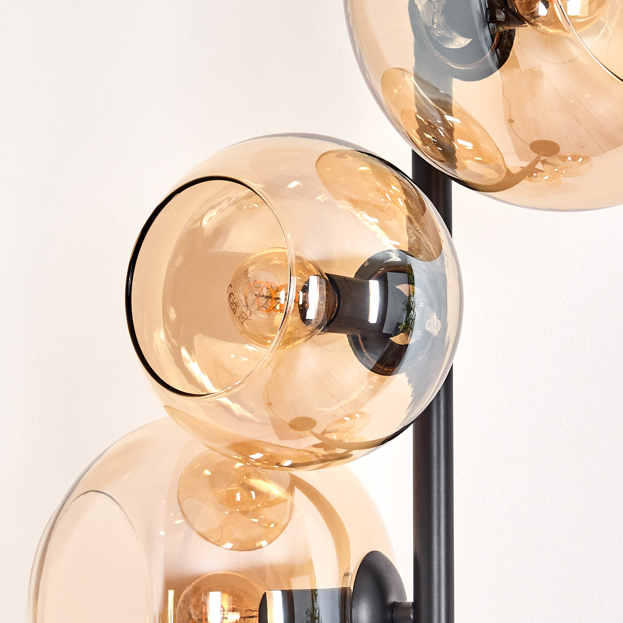 Eine moderne Lampe mit drei kugelförmigen, bernsteinfarbenen Glasschirmen. Jeder Schirm beherbergt eine Glühbirne. Die Lampe hat einen schwarzen Metallrahmen.