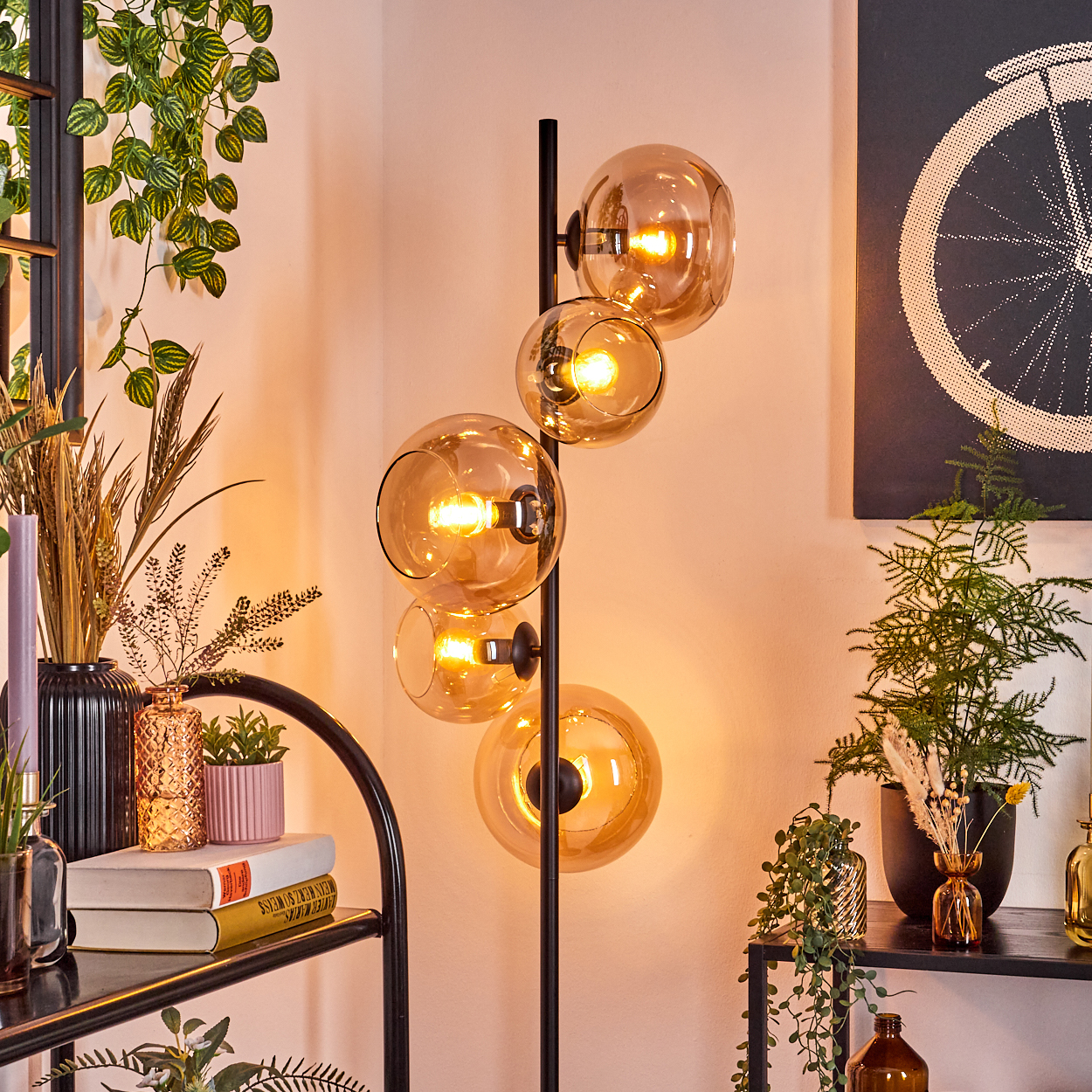 Eine moderne Stehlampe mit vier bernsteinfarbenen Glaskugeln. Um die Lampe und auf Regalen sind verschiedene Pflanzen und Dekorationsartikel angeordnet.
