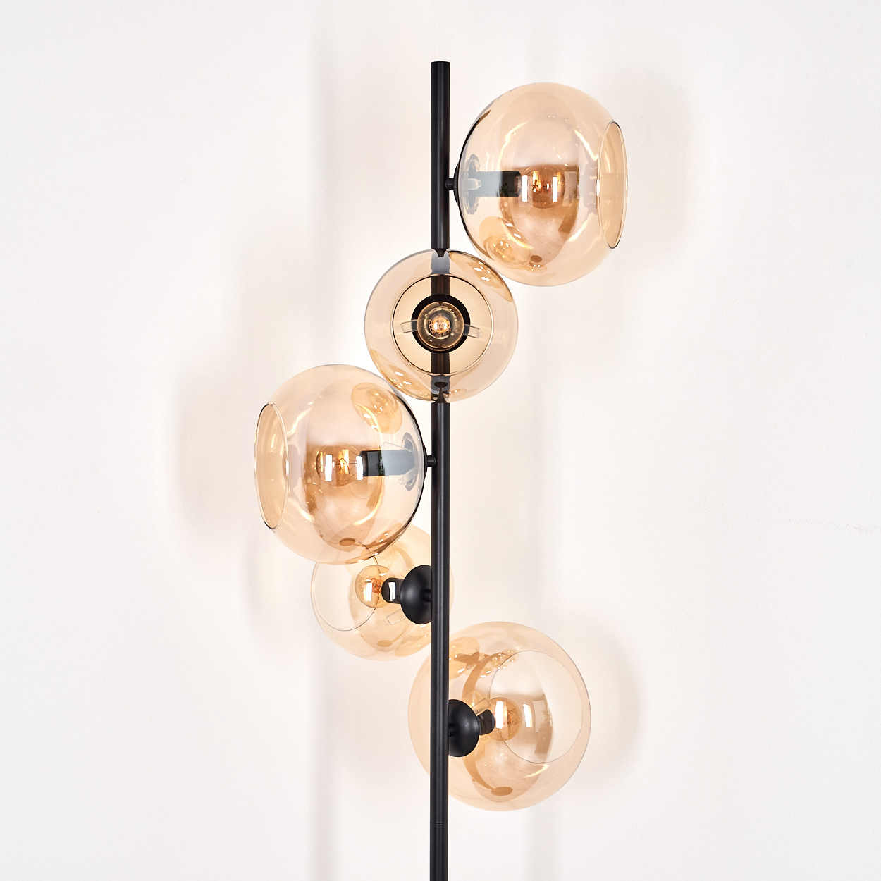 Eine moderne Stehlampe mit fünf kugelförmigen, bernsteinfarbenen Glasschirmen an einer schwarzen Metallstange vor einer weißen Wand.