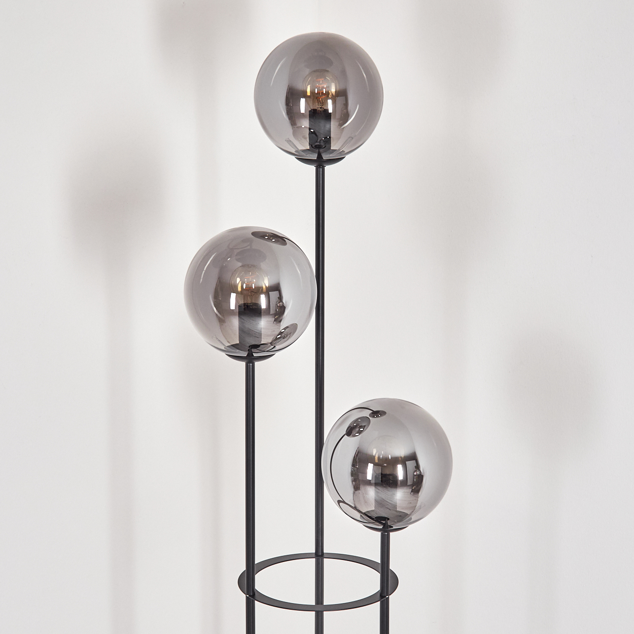 Eine moderne Stehlampe mit drei kugelförmigen Schirmen. Jeder Schirm reflektiert eine Glühbirne. Schwarzes Metallgestell. Weiße Wand im Hintergrund.