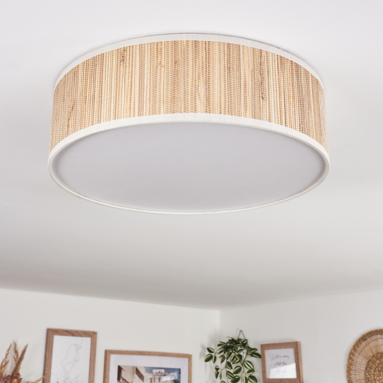 Eine runde Deckenlampe mit gewebtem Rattanschatten und weißem Diffusor. Der Hintergrund zeigt gerahmte Kunst und Pflanzen.