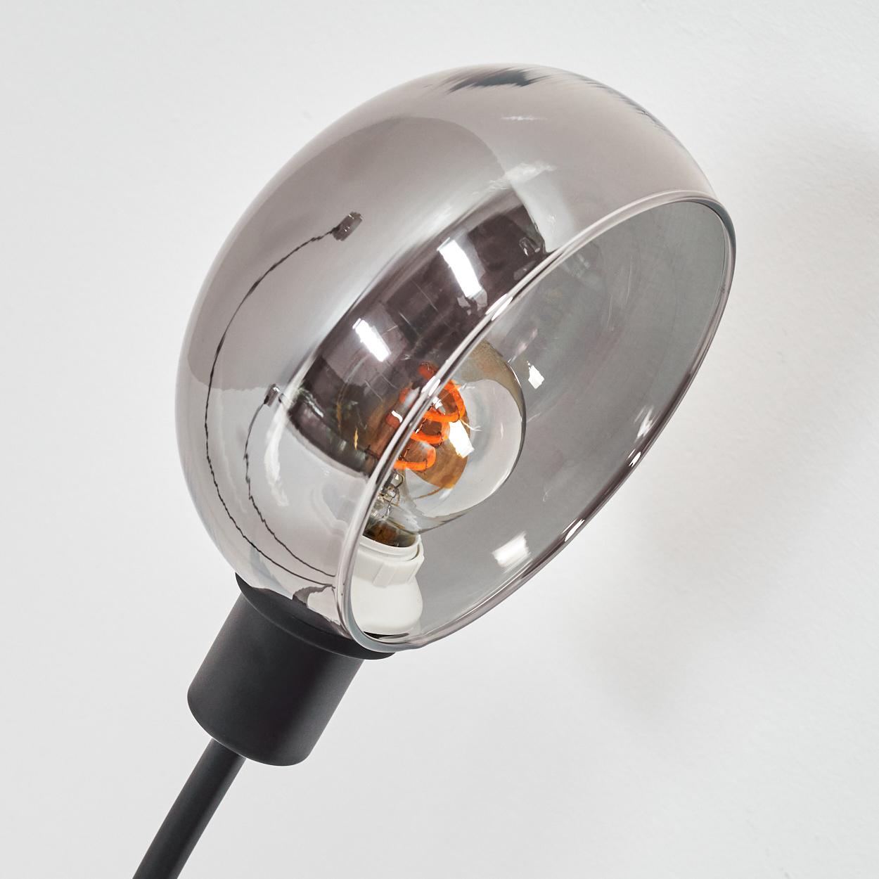 Eine moderne Lampe mit Chromschirm und sichtbarer Glühbirne. Die Lampe ist schwarz, vor einer weißen Wand.