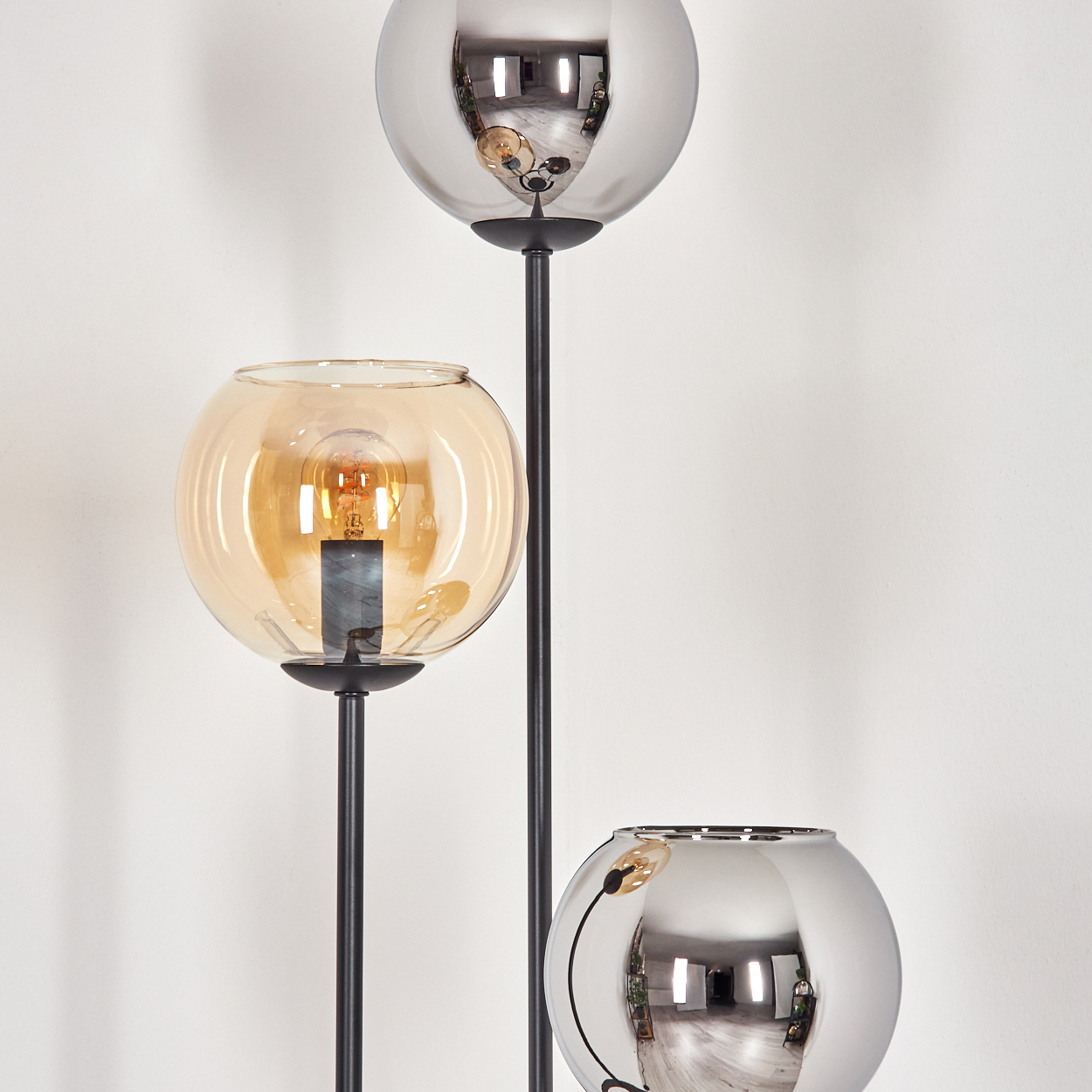 Eine moderne Stehlampe mit drei kugelförmigen Schirmen. Die oberen und unteren Schirme sind silber, der mittlere bernsteinfarben. Der Ständer ist schwarz.