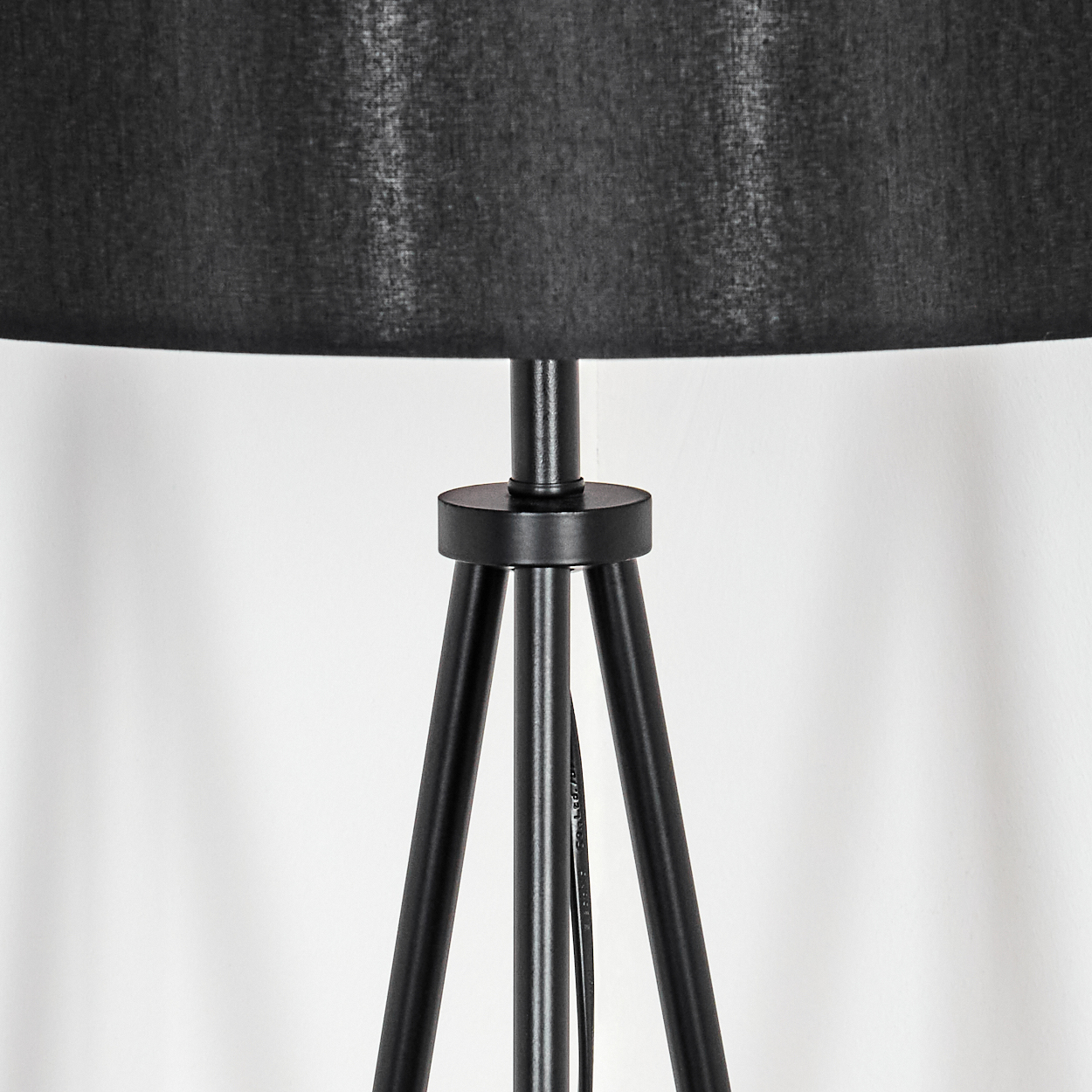 Schwarze Lampe mit Dreibein-Sockel. Der Lampenschirm ist schwarz und zylindrisch. Der Hintergrund ist eine weiße Wand.