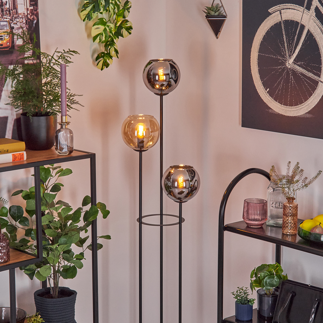 Ein Raum mit Pflanzen, Regalen und einer dreiflammigen Lampe. Ein Fahrradrad-Kunstwerk hängt an der Wand.