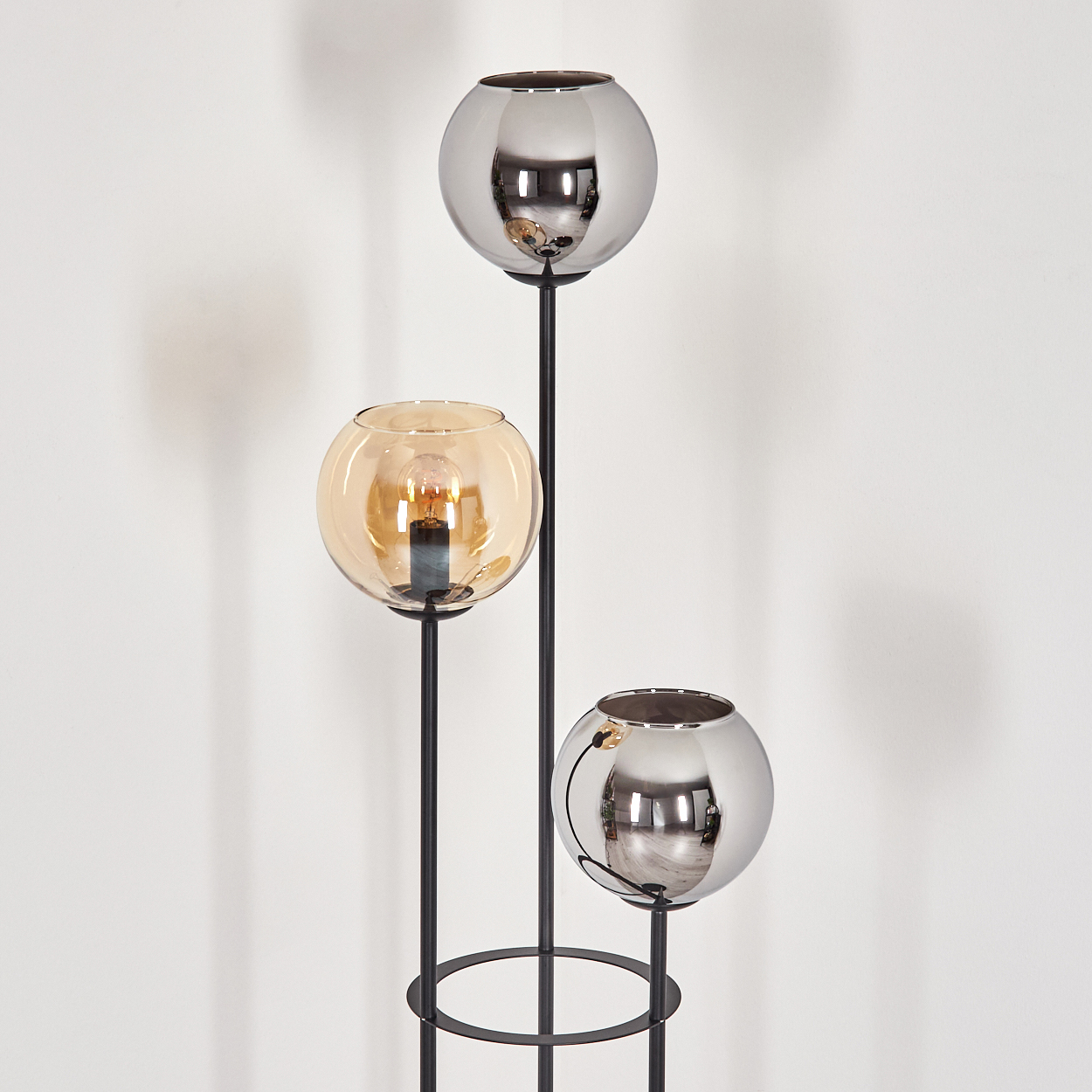 Eine moderne Stehlampe mit drei kugelförmigen Schirmen. Zwei sind verspiegelt, eine bernsteinfarben. Schwarzer Metallrahmen. Weißer Wandhintergrund.