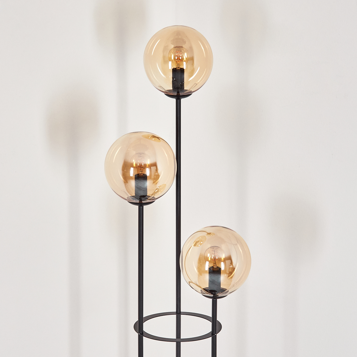 Eine moderne Stehlampe mit drei bernsteinfarbenen Glaskugelschirmen. Der schwarze Metallrahmen steht vor einer weißen Wand.