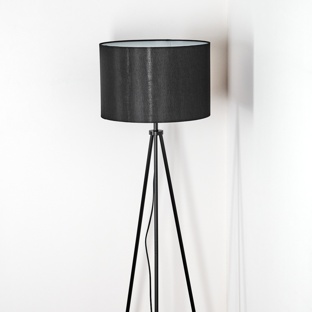 Eine schwarze Stehlampe steht in einer weißen Ecke. Die Lampe hat einen zylindrischen Schirm und ein Stativ.