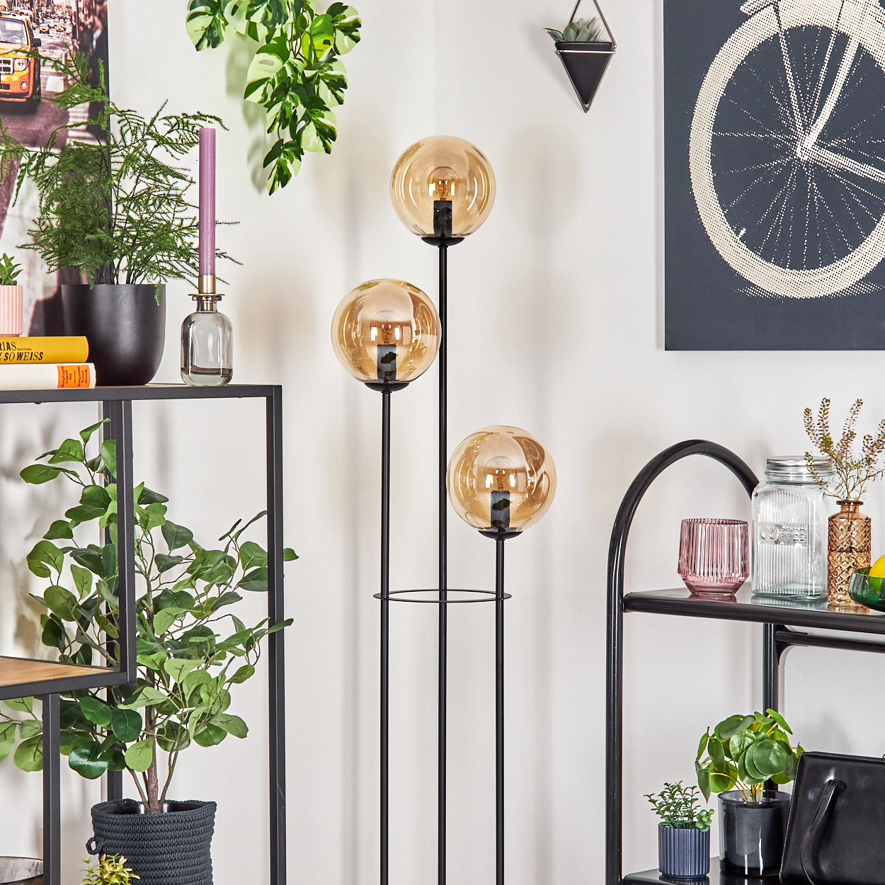 Eine Zimmerecke mit einer Stehlampe mit drei Glühbirnen, Pflanzen und Regalen. Kunstwerke und Dekorationsartikel sind sichtbar.