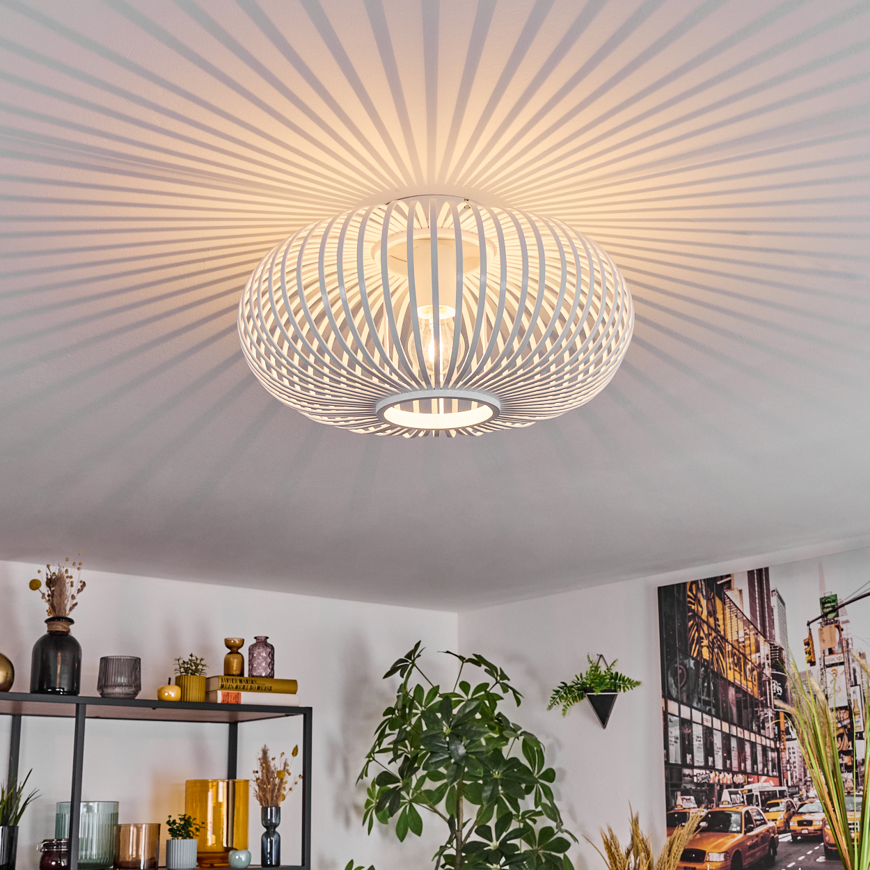 Weiße, kugelförmige Deckenlampe, die Lichtstrahlen wirft. Darunter ein Regal mit Deko, eine große Pflanze und ein Stadtbild.