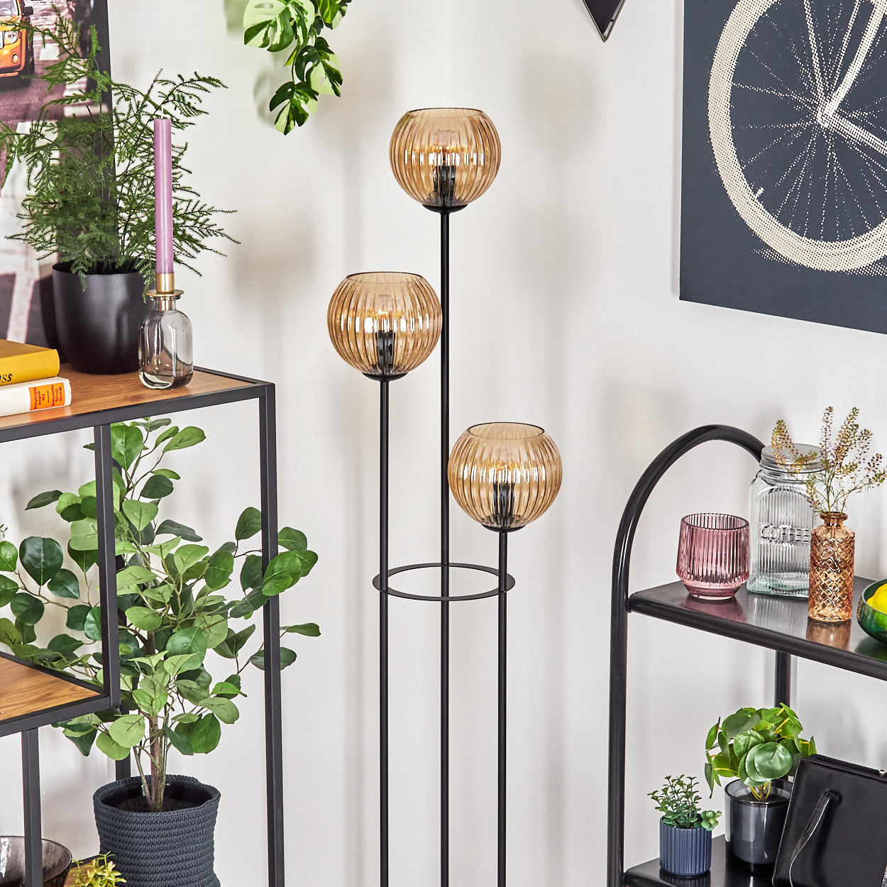 Ein Raum mit Regalen, Pflanzen und einer Stehlampe mit drei Glaskugeln. Wandkunst zeigt ein Fahrradrad. Dekorative Gegenstände sind sichtbar.