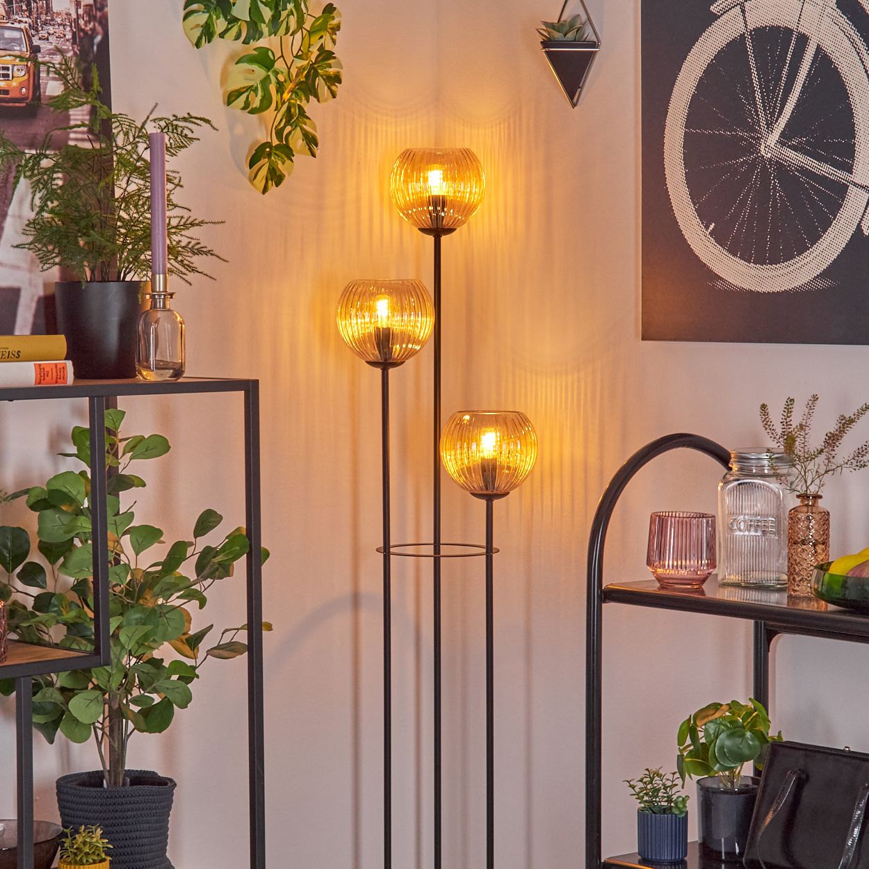 Ein modernes Zimmer mit einer Stehlampe mit drei leuchtenden Kugeln. Pflanzen, Regale und dekorative Gegenstände sind ebenfalls sichtbar.