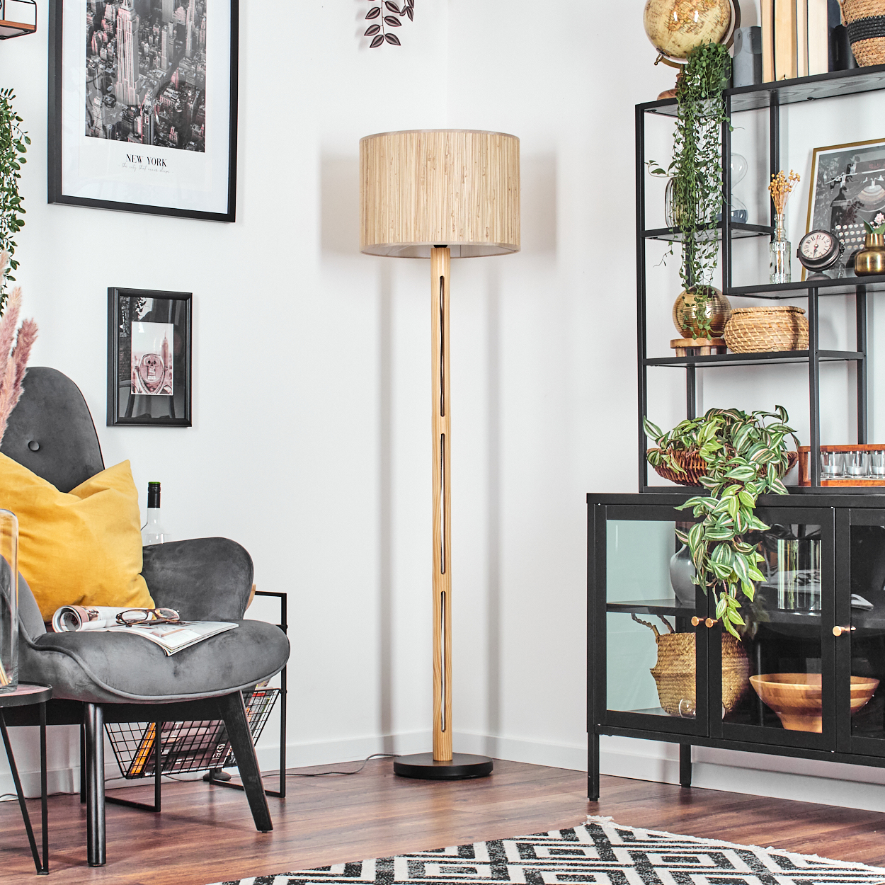 Ein Raum mit Stehlampe, Sessel, schwarzem Regal und Schrank. Bilder an der Wand und ein gemusterter Teppich.