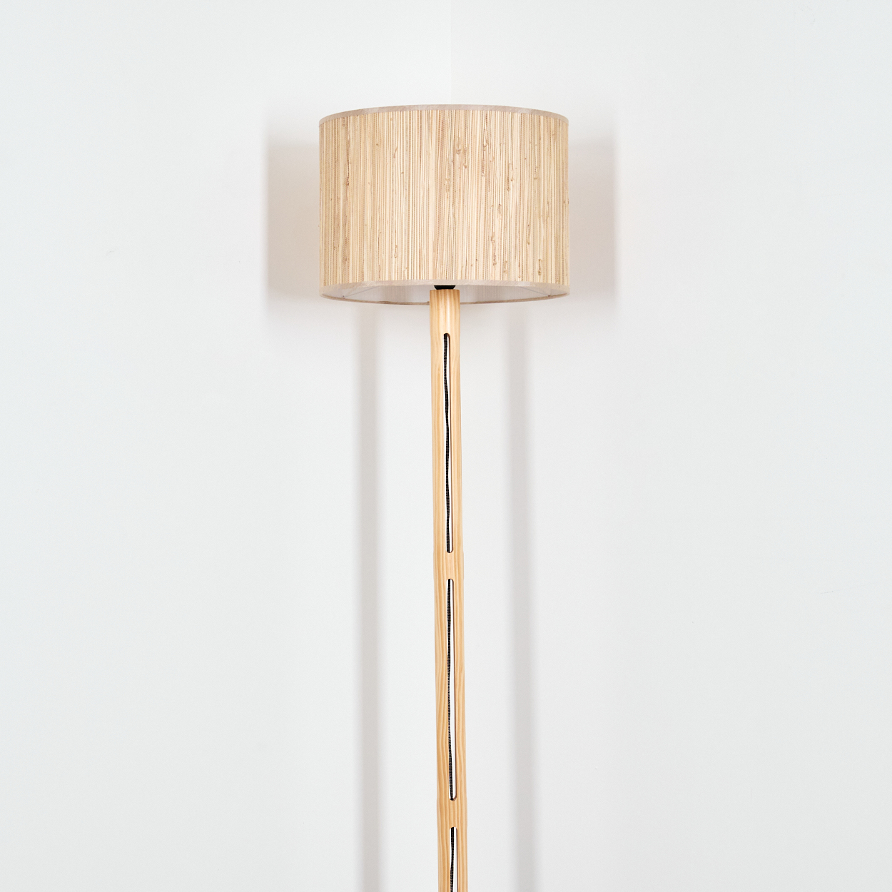 Ein beiger Lampenschirm sitzt auf einem hölzernen Lampenständer vor einer weißen Wand. Die Lampe hat einen zylindrischen Schirm und einen schlanken Sockel.