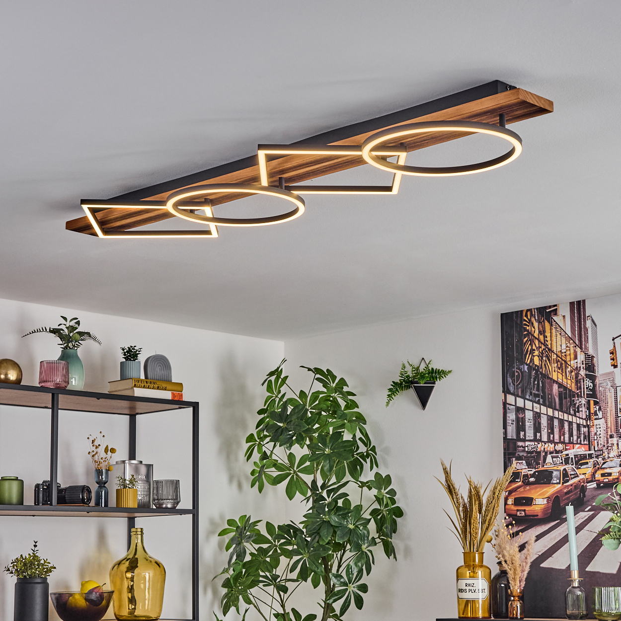 Eine moderne Deckenleuchte mit geometrischen Formen. Pflanzen und ein Regal mit Dekorationen befinden sich im Raum. Ein Stadtbild hängt an der Wand.