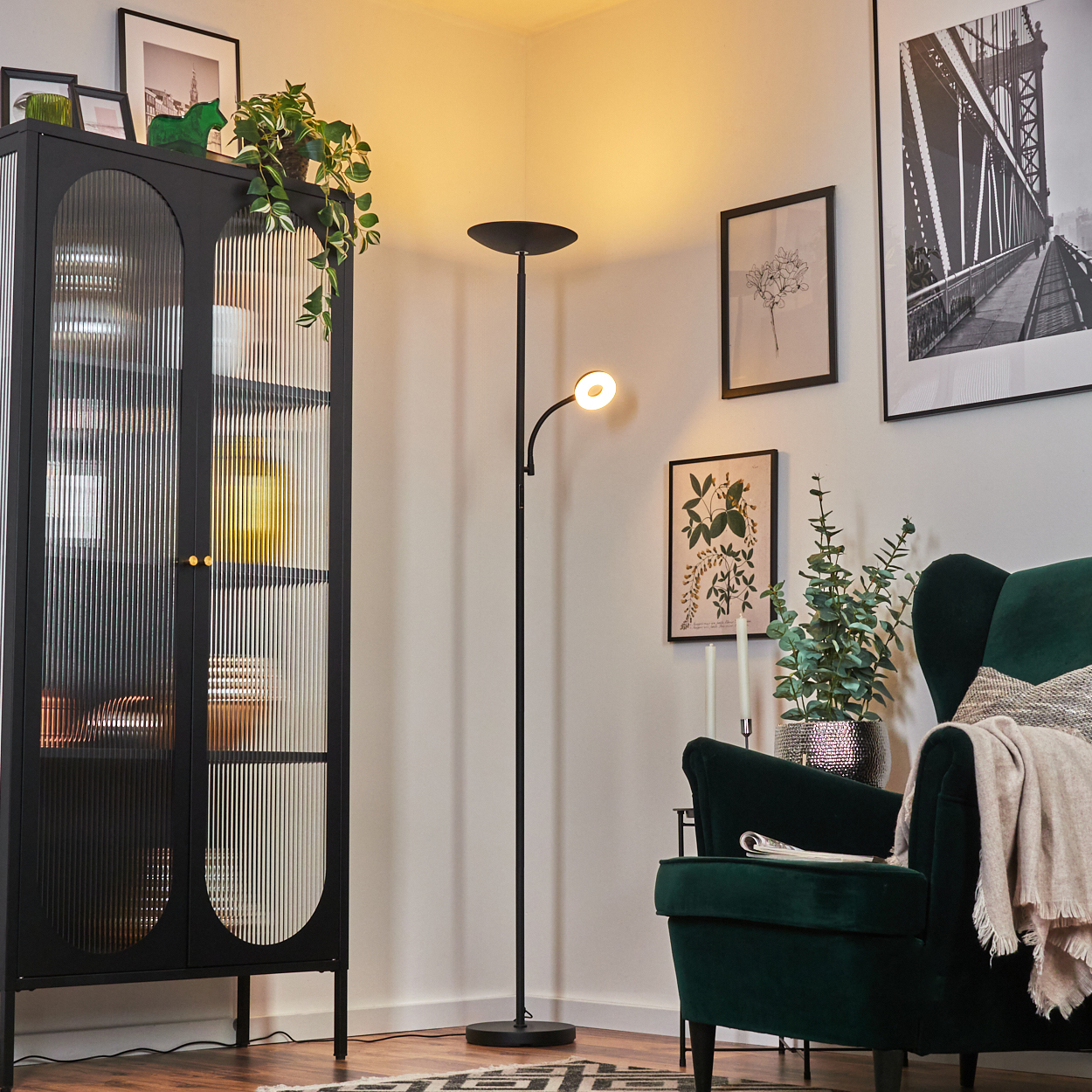Ein Raum mit schwarzem Schrank, Stehlampe und grünem Sessel. Bilder hängen an den weißen Wänden. Eine Decke liegt auf dem Stuhl.