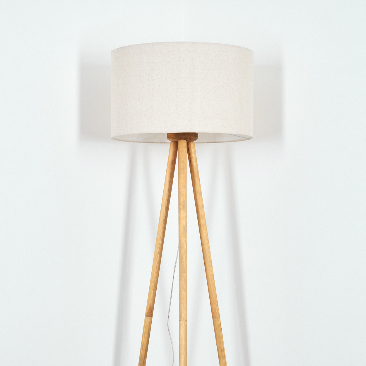 Ein beiger Lampenschirm steht auf Holzbeinen vor einer weißen Wand. Ein Kabel hängt herunter. Die Lampe wirft einen Schatten.