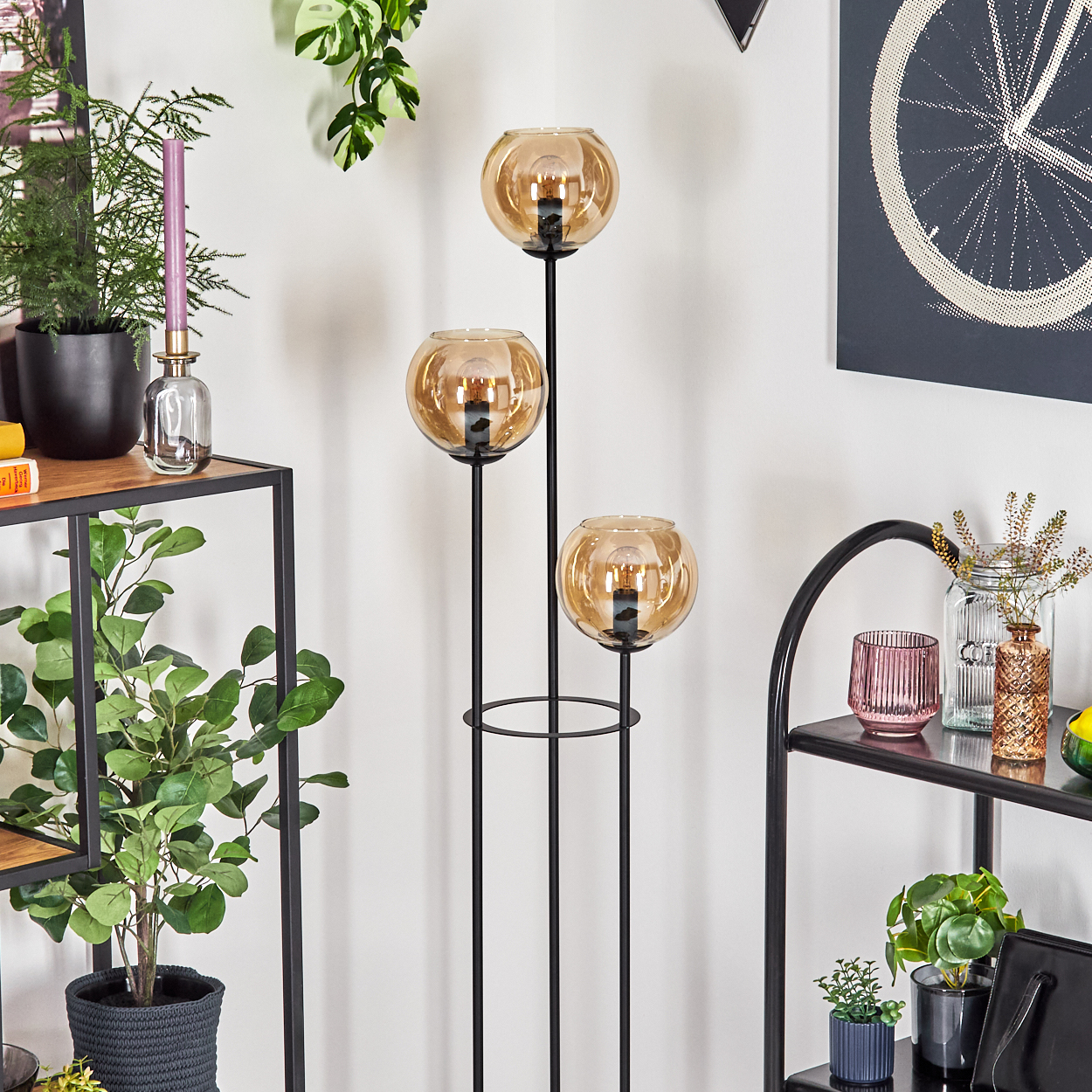 Ein modernes Interieur mit einer dreiflammigen Stehlampe, Pflanzen und dekorativen Gegenständen auf Regalen vor einer weißen Wand.