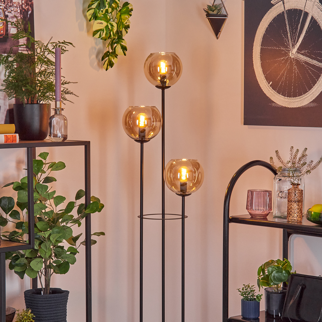 Ein modernes Wohnzimmer mit einer schwarzen Metall-Stehlampe mit drei Kugelleuchten. Pflanzen und Regale vervollständigen die Szene.