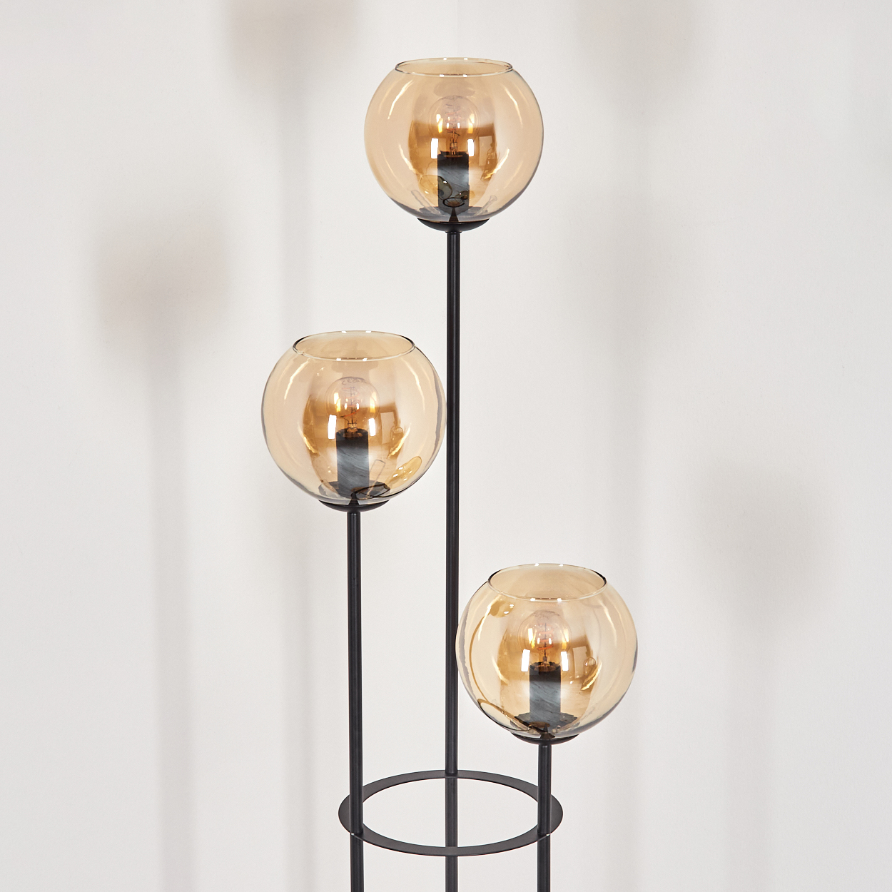 Eine moderne Stehlampe mit drei kugelförmigen, bernsteinfarbenen Glasschirmen. Die Lampe hat einen schwarzen Metallrahmen. Der Hintergrund ist eine weiße Wand.