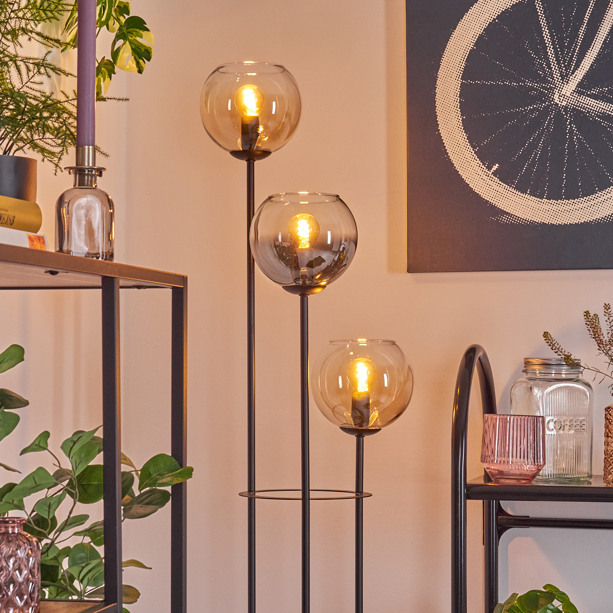 Eine Stehlampe mit drei Glaskugelschirmen. Der Raum hat ein Regal mit Pflanzen, einer Kerze, einem Radbild und einem Kaffeebehälter.