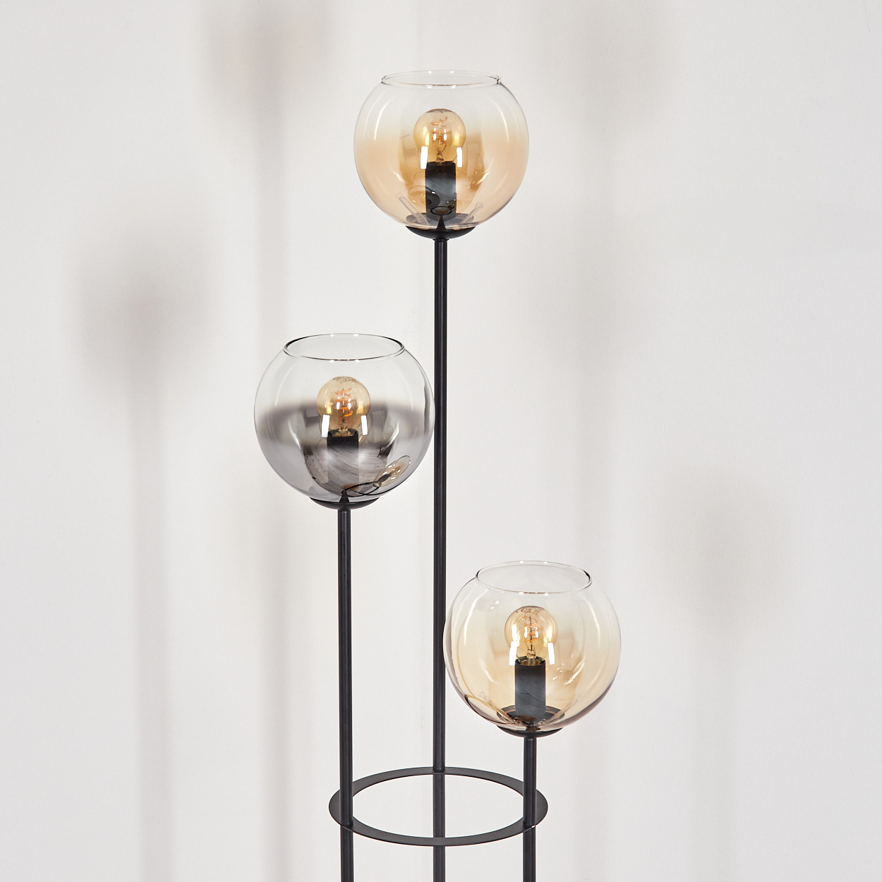 Eine moderne Stehlampe mit drei Glaskugelschirmen. Jede enthält eine Glühbirne. Der Ständer ist schwarz, vor einer weißen Wand.