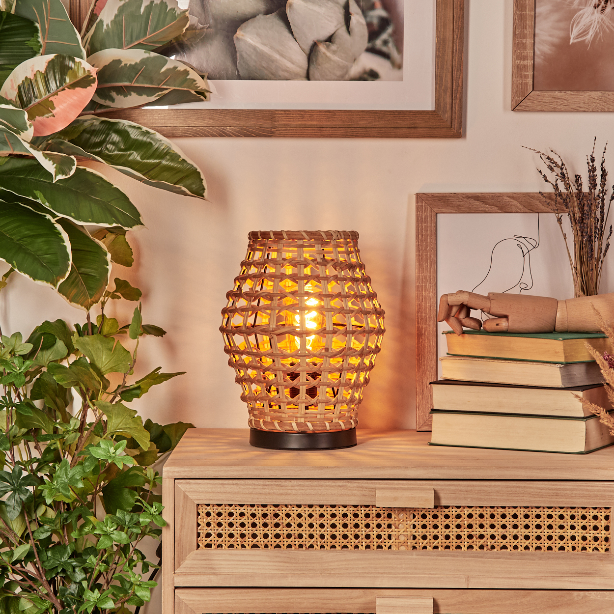Eine gewebte Lampe mit einer brennenden Glühbirne steht auf einem Holzschrank. Pflanzen und gerahmte Bilder sind im Hintergrund.