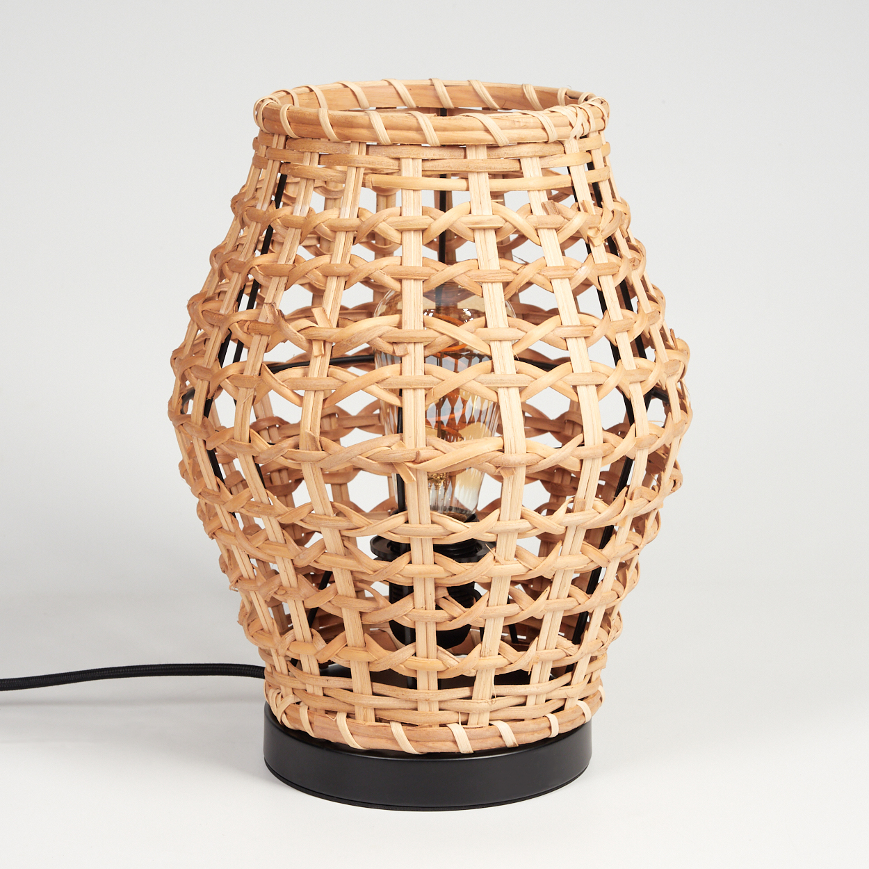Eine gewebte Rattanlampe mit einer freiliegenden Glühbirne. Sie hat einen schwarzen Sockel und ein schwarzes Netzkabel vor weißem Hintergrund.