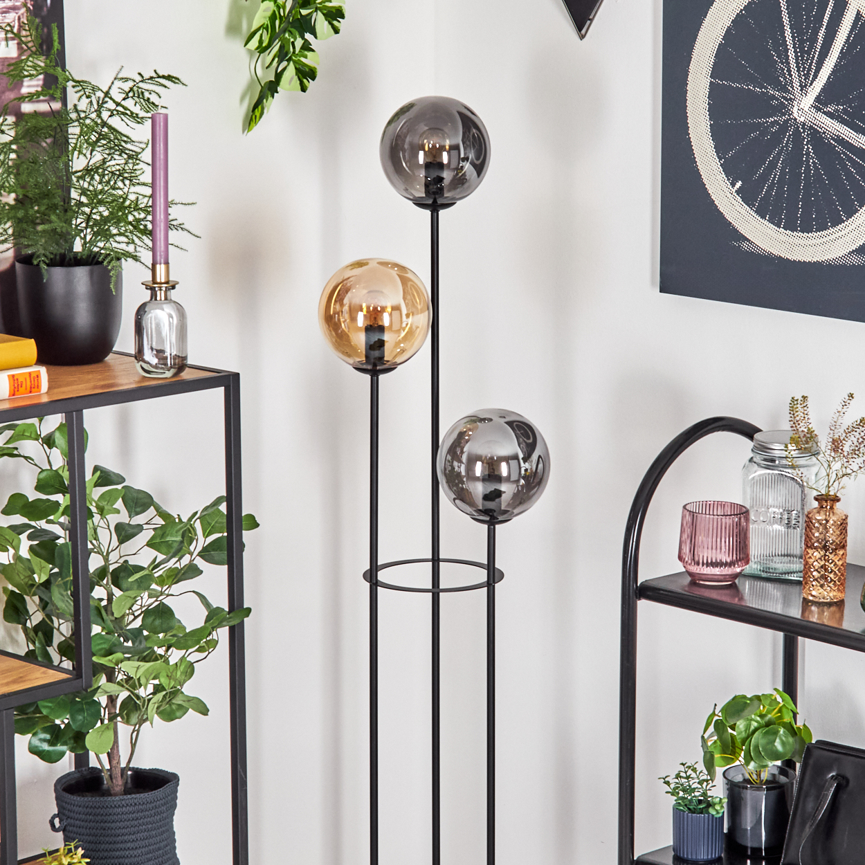 Stehlampe mit drei Kugellampenschirmen. Regale mit Pflanzen, Kerze und Deko. Fahrradrad-Kunst an der Wand.
