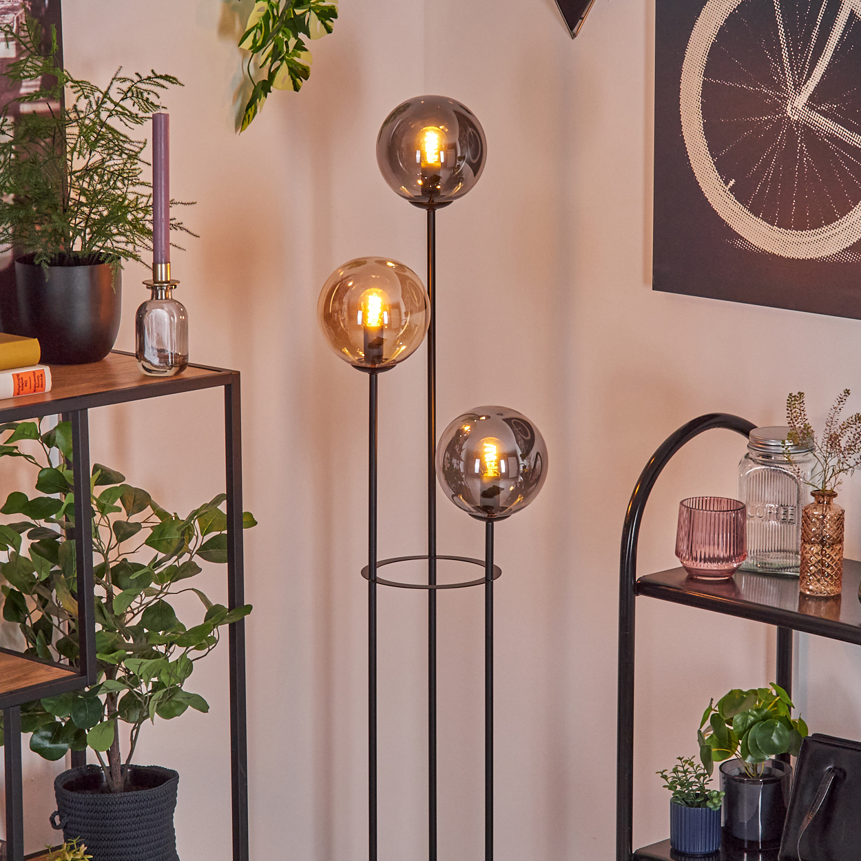 Ein Raum mit einer Stehlampe mit drei Kugelleuchten. Es gibt Pflanzen, Regale und eine dekorative Wandkunst.