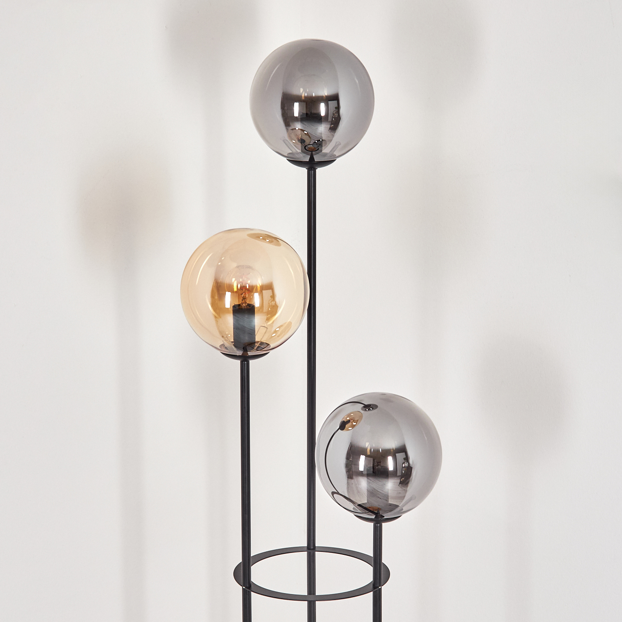 Eine moderne Stehlampe mit drei kugelförmigen Schirmen. Zwei sind silberfarben, einer ist bernsteinfarben. Der Ständer ist aus schwarzem Metall vor einer weißen Wand.