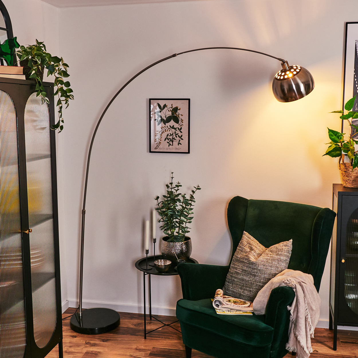 Ein Zimmer mit grünem Sessel, Stehlampe und schwarzem Schrank. Pflanzen und Dekoration sind ebenfalls vorhanden. Ein Bild hängt an der Wand.