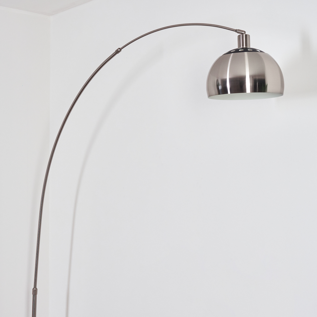 Eine moderne Bogenstehlampe mit silbernem Kuppelschirm. Der gebogene Arm der Lampe erstreckt sich vom Sockel zum Schirm, vor einer weißen Wand.