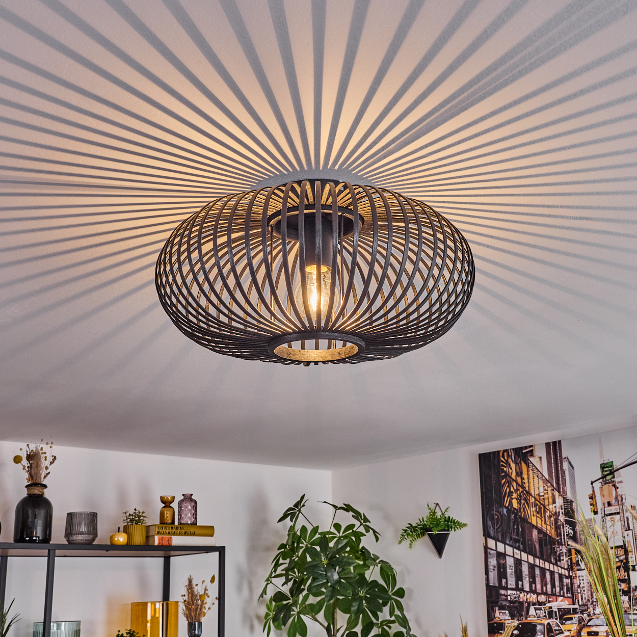 Eine moderne schwarze Deckenlampe wirft einen Schatten auf die weiße Decke. Der Raum enthält Pflanzen, Regale und ein Stadtbildgemälde.